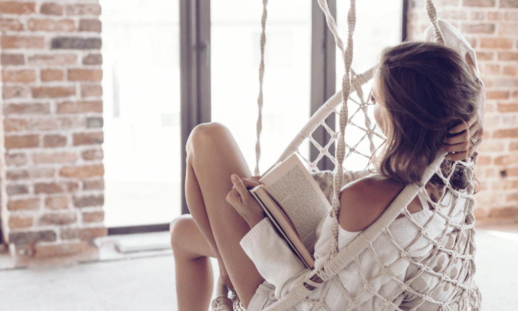 A woman in her 40s is reading a book while resting in a white fabric hammock in her apartment living room. The woman is wearing a comfortable outfit and has her hair comfortably lose. The windows are open, letting in natural light. The brick walls are painted a light color. The headline of the blog post is "The Best Foods to Eat for Hormone Health After 40."